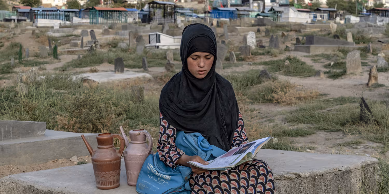 A girl sitting on a stone reading a book with a UN Women bag next to her. A number of ordinary-looking flats in the background.