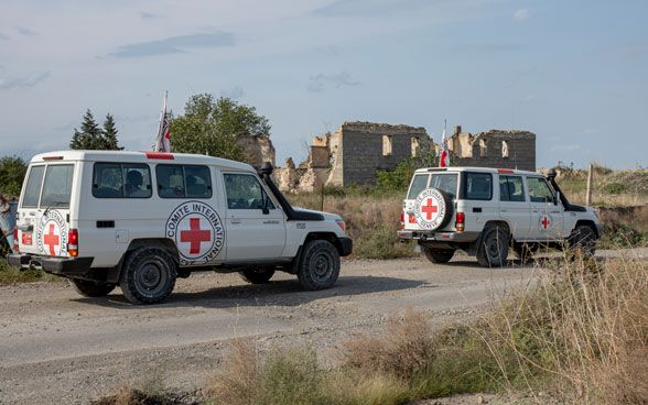 Two ICRC vehicles on the move with destroyed houses in the background.