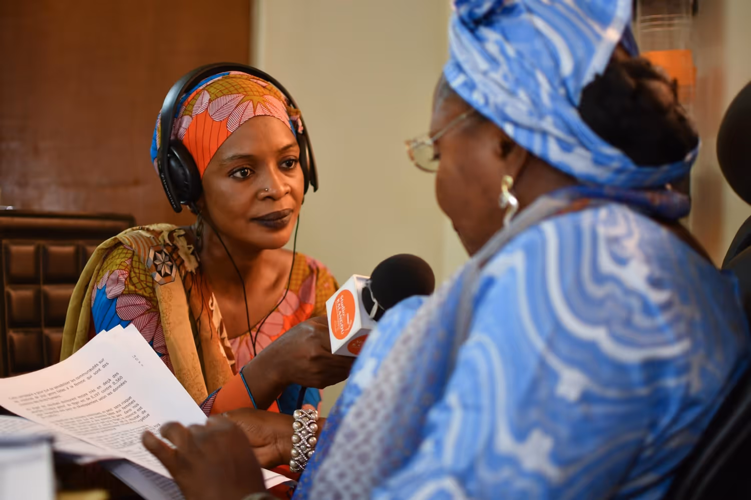 An African woman holds a microphone as she interviews another woman in a radio studio.