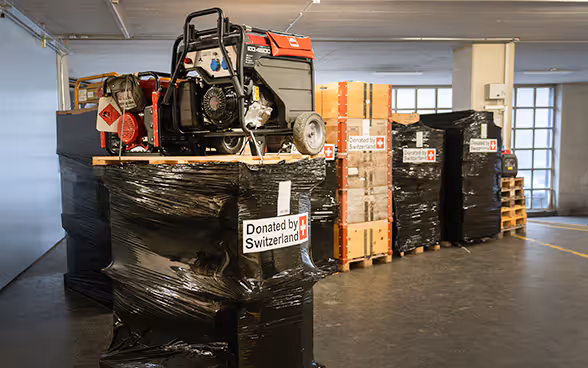 In a warehouse, a box with a picture of a mobile heater on a pallet is in the foreground. In the background are more pallets on which packed electricity generators are placed.