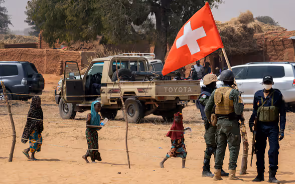 Some soldiers stand near a car. One person holds up a Swiss flag.