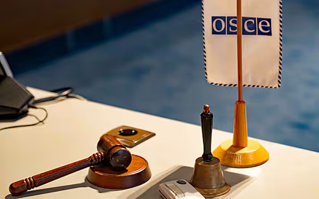 A wooden hammer, a bell and a small flag with OSCE written on it are arranged on a table.