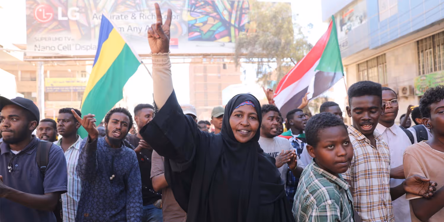A woman standing in a group of people, making the victory sign.