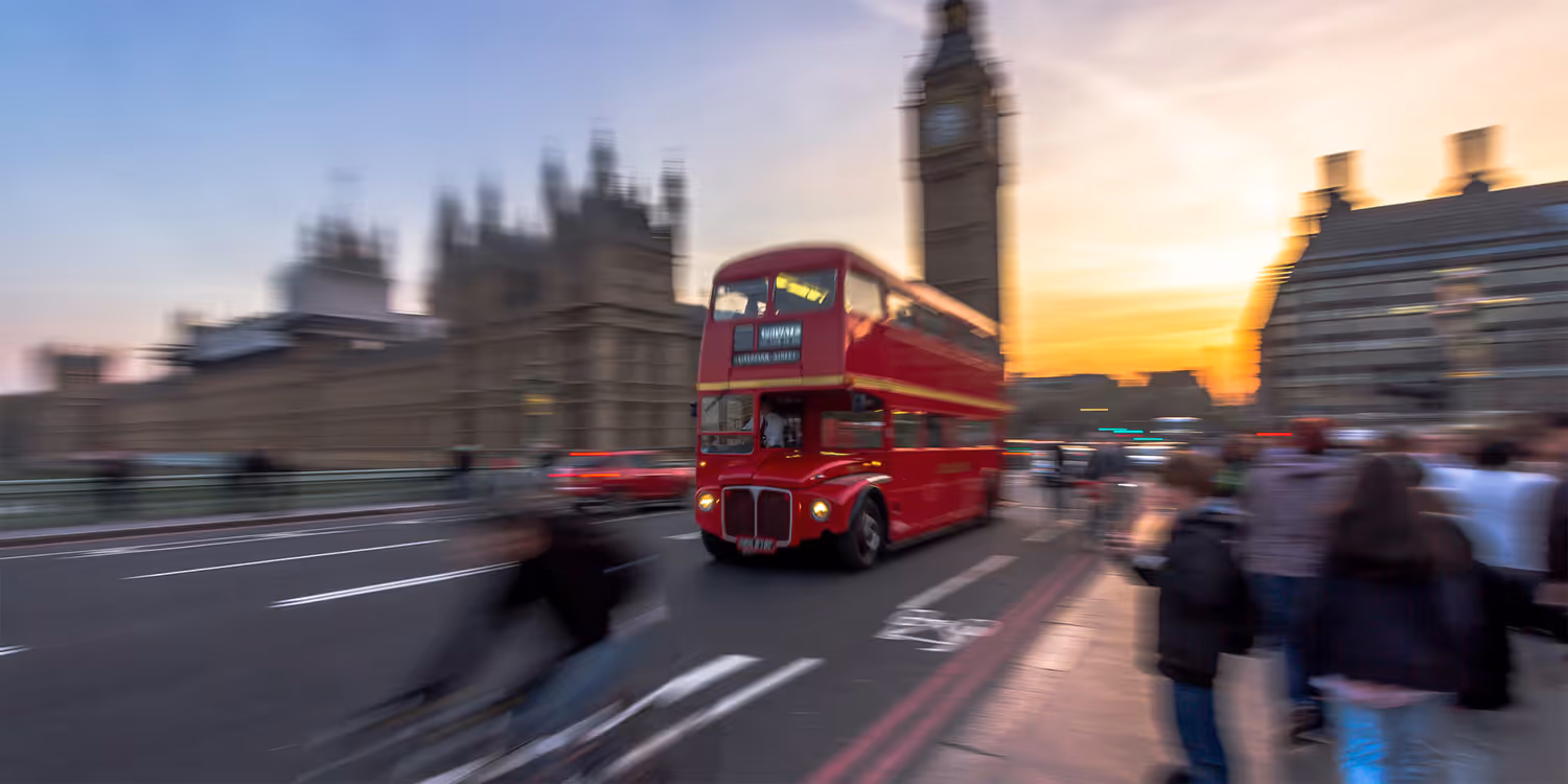 A red double-decker bus, typical of London, drives past Big Ben over Westminster Bridge into the sunset.