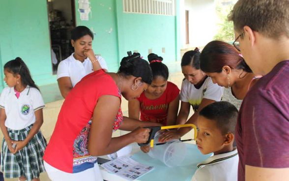 A young woman sawing through a water filter on an outdoor table. Two other women are helping her. Several people are standing watching.