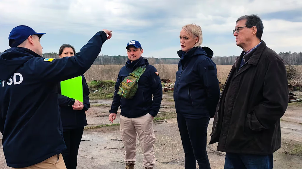 A man gives explanations to a small group of people and points to a plot of land on the edge of a forest.