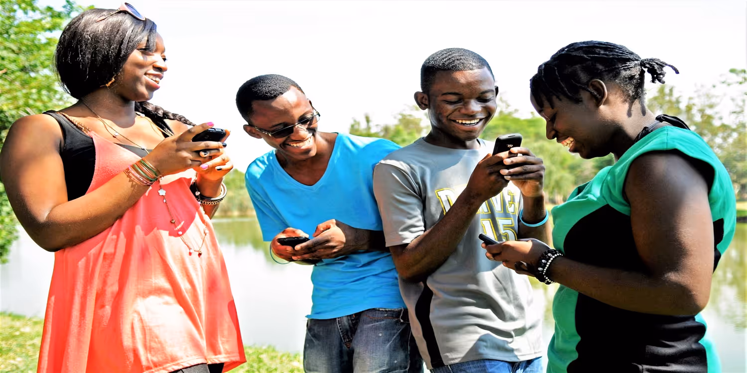 Four young people in the foreground smile and look at their mobile phone screens.