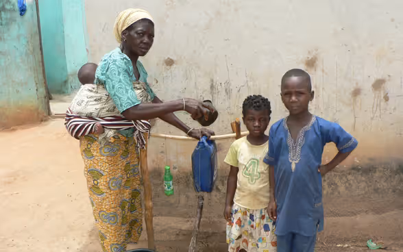 A woman with an infant on her back shows two children how to fill the makeshift handwashing device with water in front of a hut.
