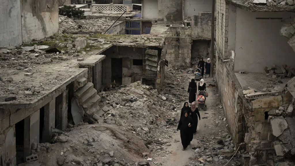 Women walk with prams through the rubble of a street with collapsed houses. 