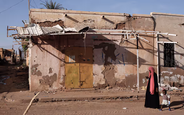 Une femme tient un enfant par la main et marche devant un bâtiment en ruines. 