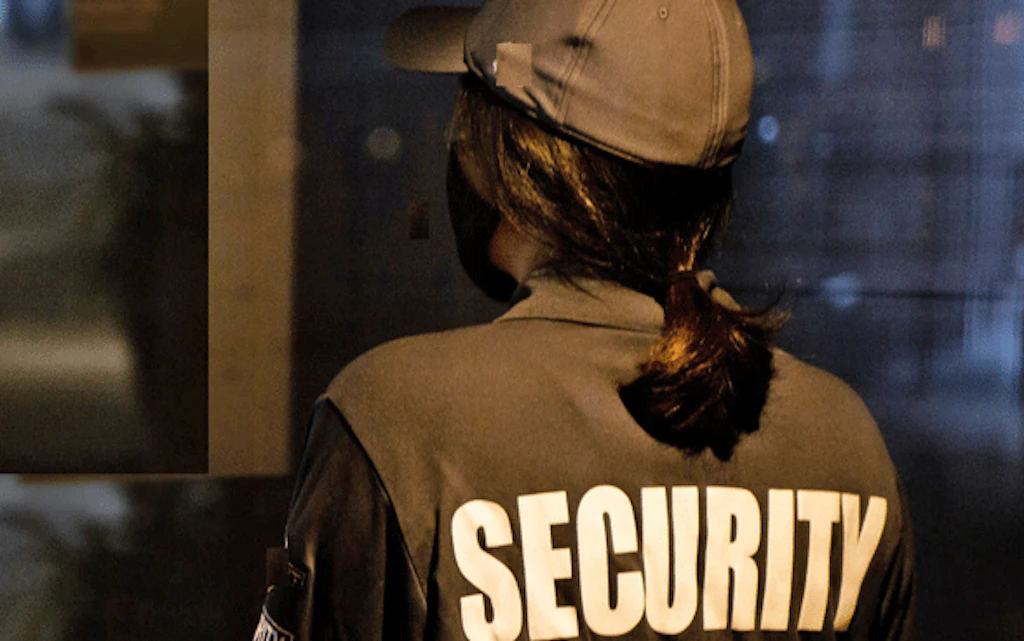 Une femme de dos, avec un uniforme portant l’inscription « Security ».  