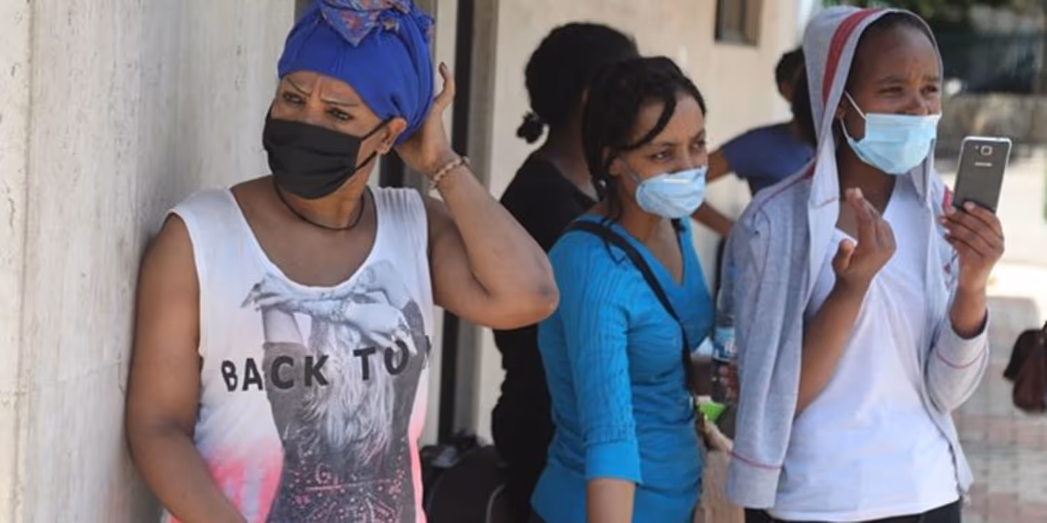 Trois femmes avec des masques attendent à l'extérieur d'un bâtiment. L'une d'entre ellesux tient un téléphone.