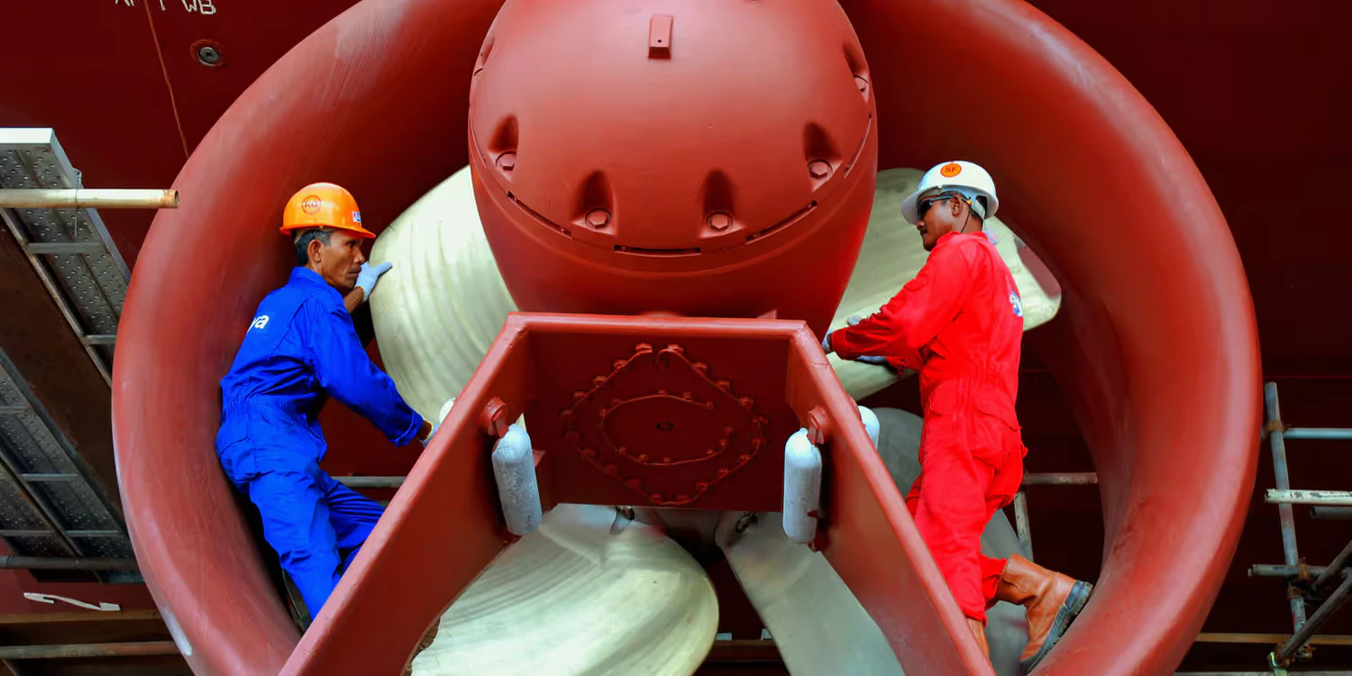 Deux travailleurs en combinaison bleu et rouge travaillent sur un réacteur dans un hangar.