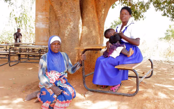Une femme est assise par terre, à l’ombre d’un arbre. À ses côtés, une autre femme, assise sur une chaise d’école, berce un bébé dans ses bras.