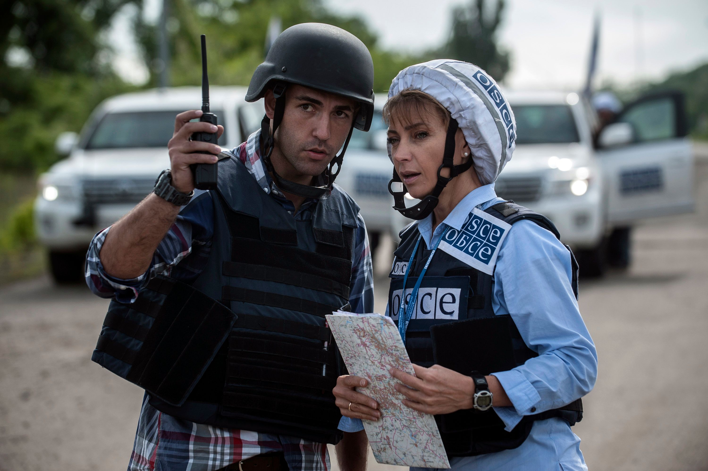 Un homme, muni d’un talkie-walkie, donne des indications à une femme, qui tient une carte à la main. Tous deux sont équipés d’un casque et de vêtements de protection. La femme porte un gilet portant pour inscription « OSCE ».