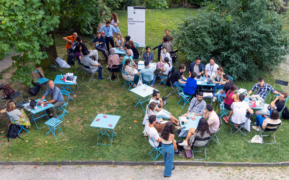 Dans une prairie, des jeunes gens discutent assis autour de tables.