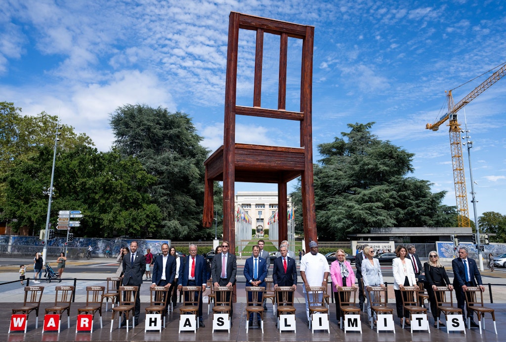 Photo de la cérémonie commémorative du 75e anniversaire des Conventions de Genève, sur la Place des Nations à Genève. Environ vingt personnes se tiennent devant la sculpture du Broken Chair et s’appuient sur des chaises brunes portant l’inscription « War has limits ».