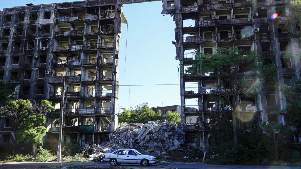 Une voiture blanche garée devant des bâtiments civils éventrés par un bombardement et laissant transparaître le ciel bleu.