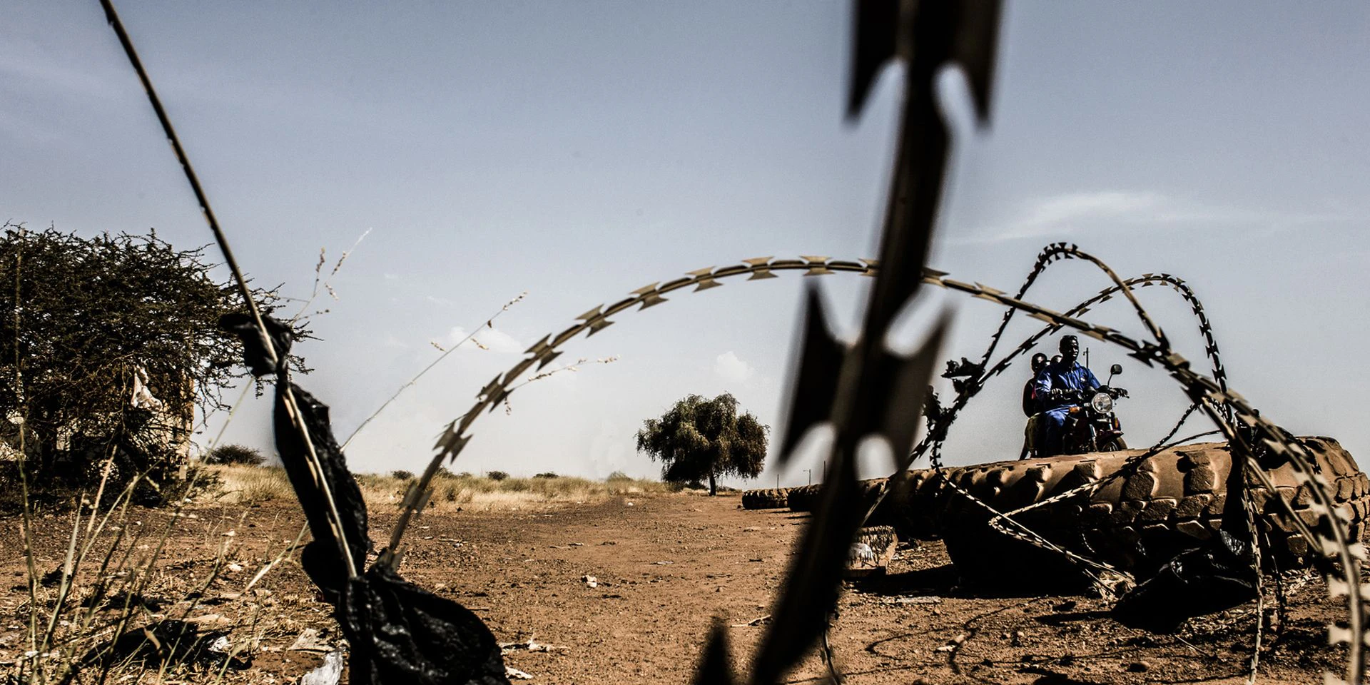 Un fil de fer barbelé et des pneus sur une route sablonneuse en Afrique. En arrière-plan, deux personnes à moto.