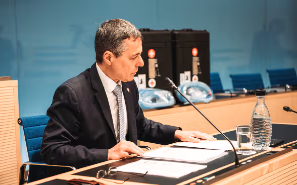 Le conseiller fédéral Cassis est assis à un bureau, les dossiers sont sur la table devant lui. En arrière-plan, vous pouvez voir les deux valises avec les respirateurs.