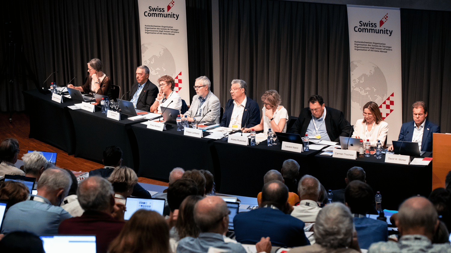 Des hommes et des femmes sont assis en rang derrière une table, au milieu se tient Filippo Lombardi. 