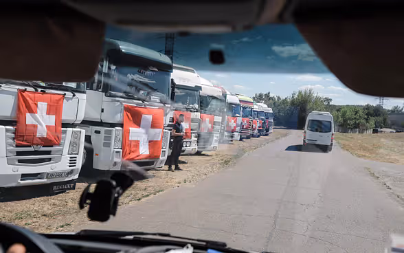 Dix camions sur lesquels s’affichent des drapeaux suisses sont alignés dans un champ en Ukraine.