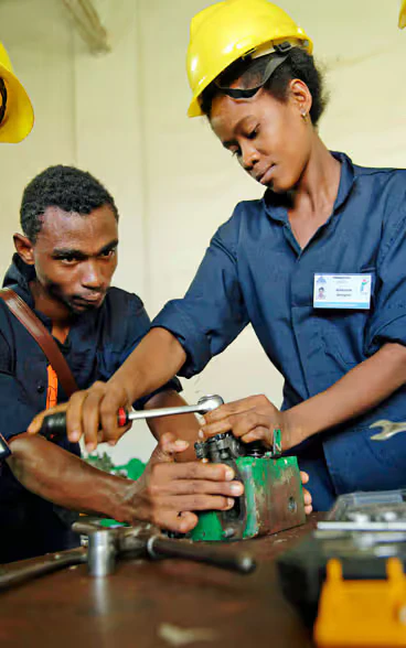 Une formatrice, munie d’un casque jaune, aide un apprenti dans un atelier.