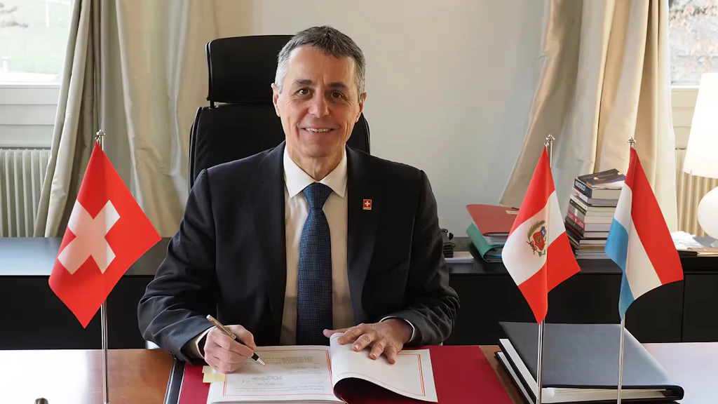 Il consigliere federale Ignazio Cassis firma un documento. Al suo fianco le bandiere della Svizzera (a destra), del Perù e del Lussemburgo (a sinistra).