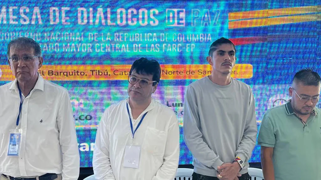 Quattro uomini dall'aria seria sono in piedi davanti a una parete che raffigura il logo del Dialogo di Pace.