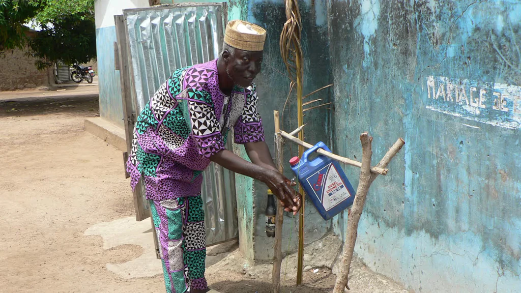 Benin: un uomo davanti a una casa si lava accuratamente le mani a un rubinetto improvvisato.