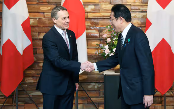 Fumio Kishida e Ignazio Cassis si stringono la mano. Sullo sfondo si vedono le bandiere del Giappone e della Svizzera. 