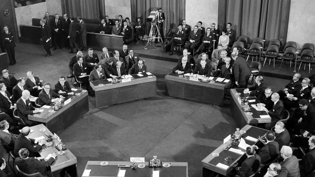 Immagine in bianco e nero: in una sala conferenze sono disposti in cerchio sette tavoli, ai quali siedono numerosi uomini.