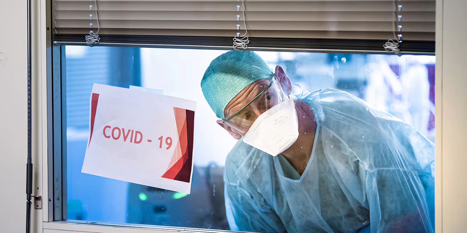 Dal reparto di terapia intensiva, un membro del personale infermieristico guarda attraverso una finestra su cui è apposta la scritta “Covid-19”.