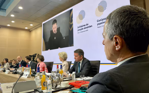 Il consigliere federale Ignazio Cassis siede a un tavolo e ascolta il presidente ucraino Zelensky, visibile in video su uno schermo.