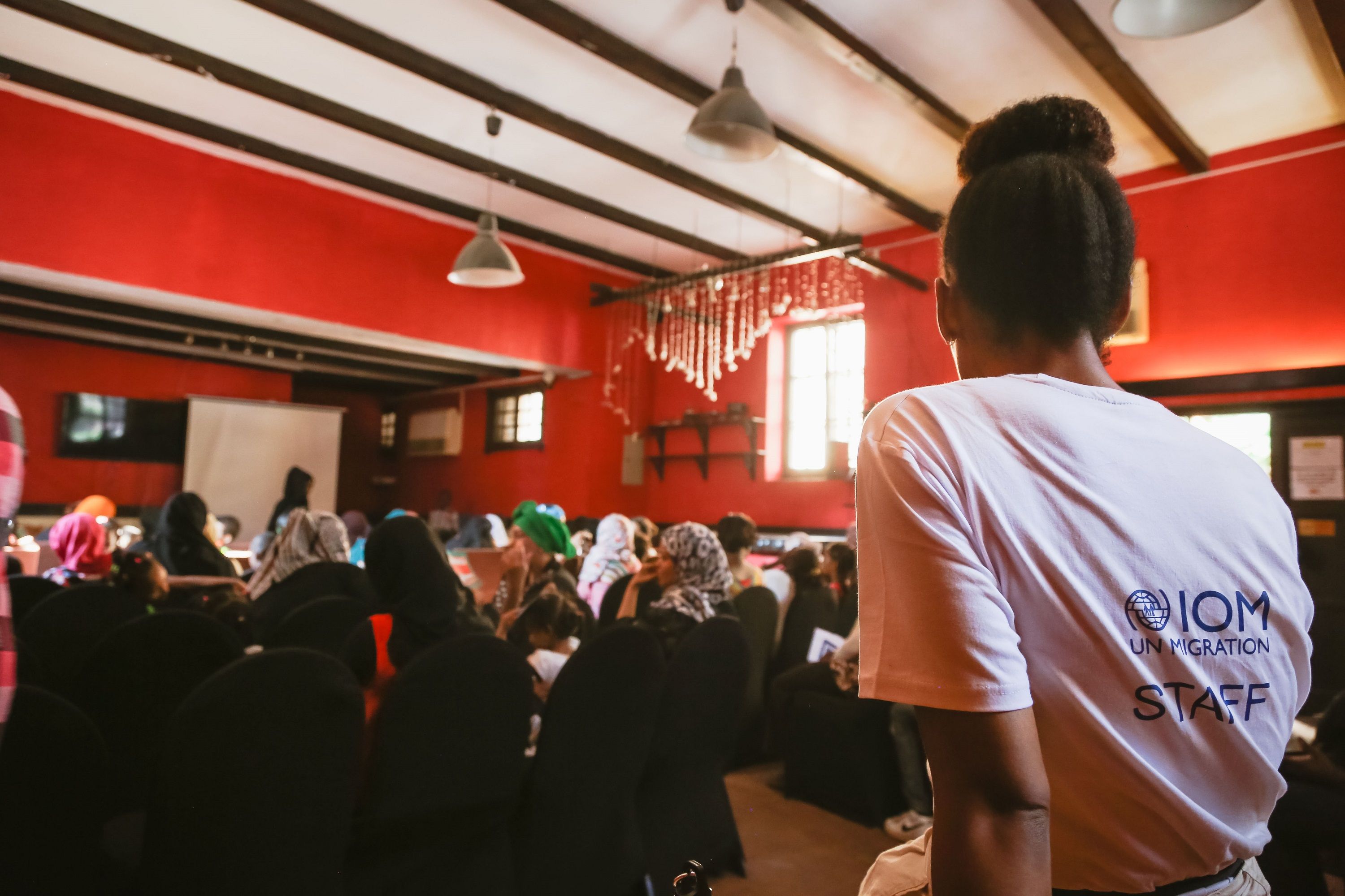Alcune persone sedute in file in una stanza guardano uno schermo, in primo piano una volontaria dell’OIM da dietro.