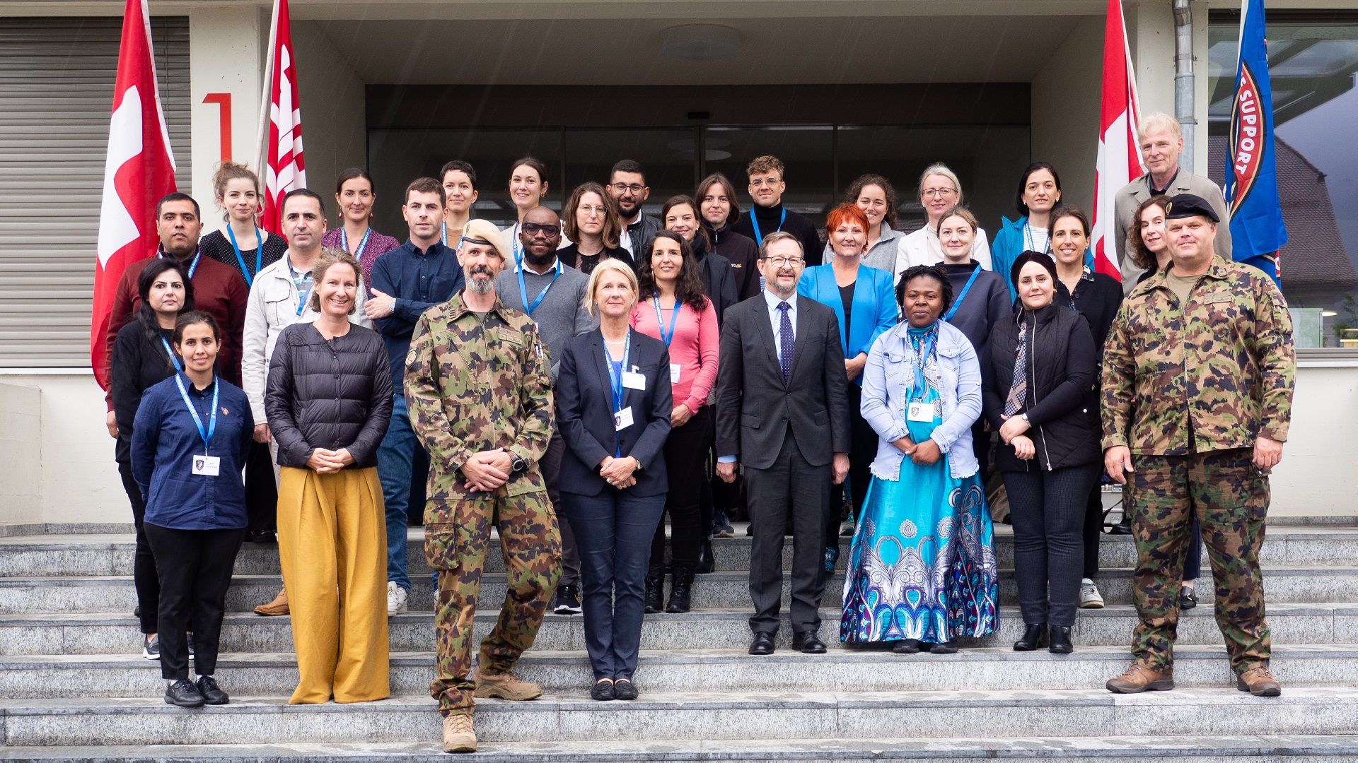 Partecipanti al corso in piedi sulla scala del centro di formazione SWISSINT di Stans. Due persone sono in uniforme militare. L’obiettivo inquadra uomini e donne che sorridono.