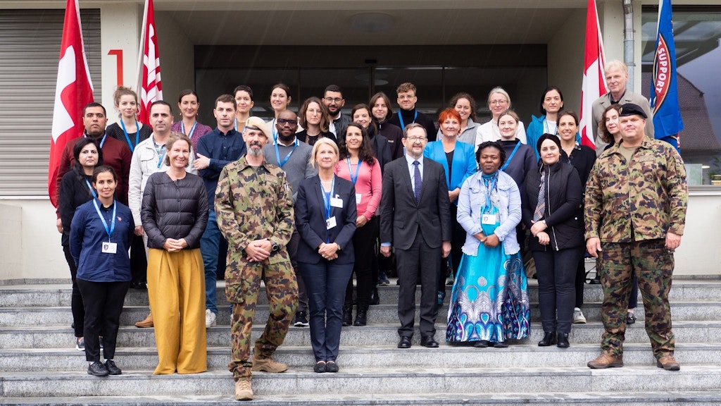 Partecipanti al corso in piedi sulla scala del centro di formazione SWISSINT di Stans. Due persone sono in uniforme militare. L’obiettivo inquadra uomini e donne che sorridono.