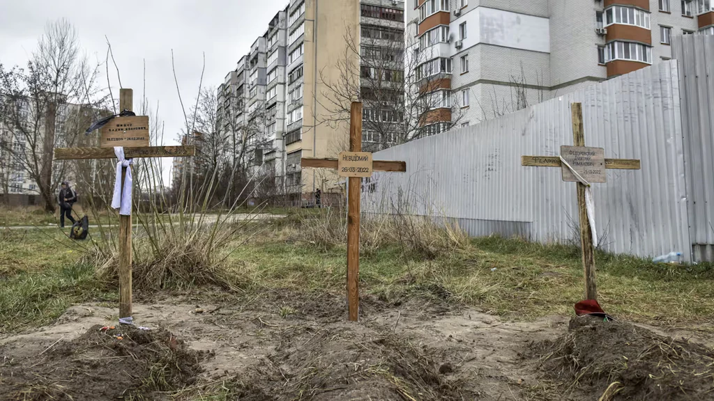 Tre tombe con croci di legno e nomi sono visibili in primo piano. Sullo sfondo delle case grandi.