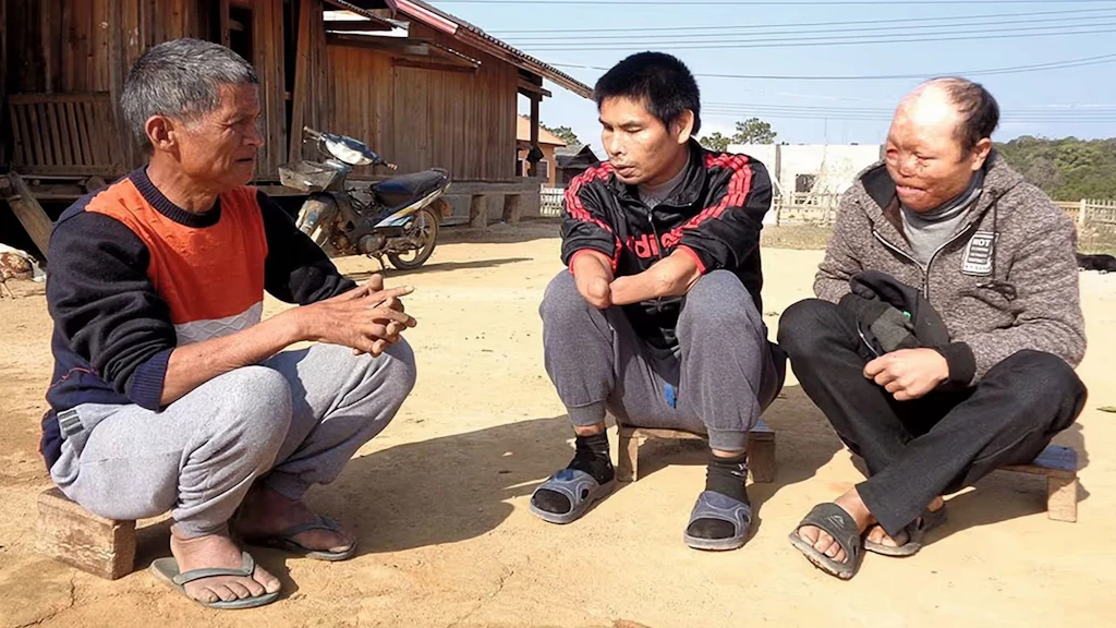 Tre vittime laotiane di munizioni a grappolo sono sedute a terra davanti a una fattoria.