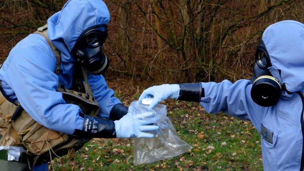 Ispettori dell’OPCW con tute protettive blu e maschere antigas esaminano un oggetto sospetto a terra durante un’esercitazione all’aperto.