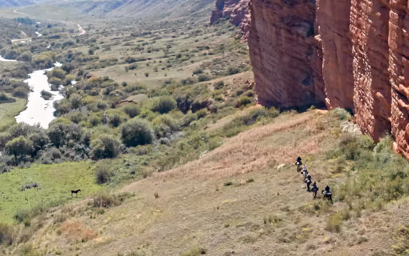 Cinque persone a cavallo alla base di un costone roccioso in una valle remota.