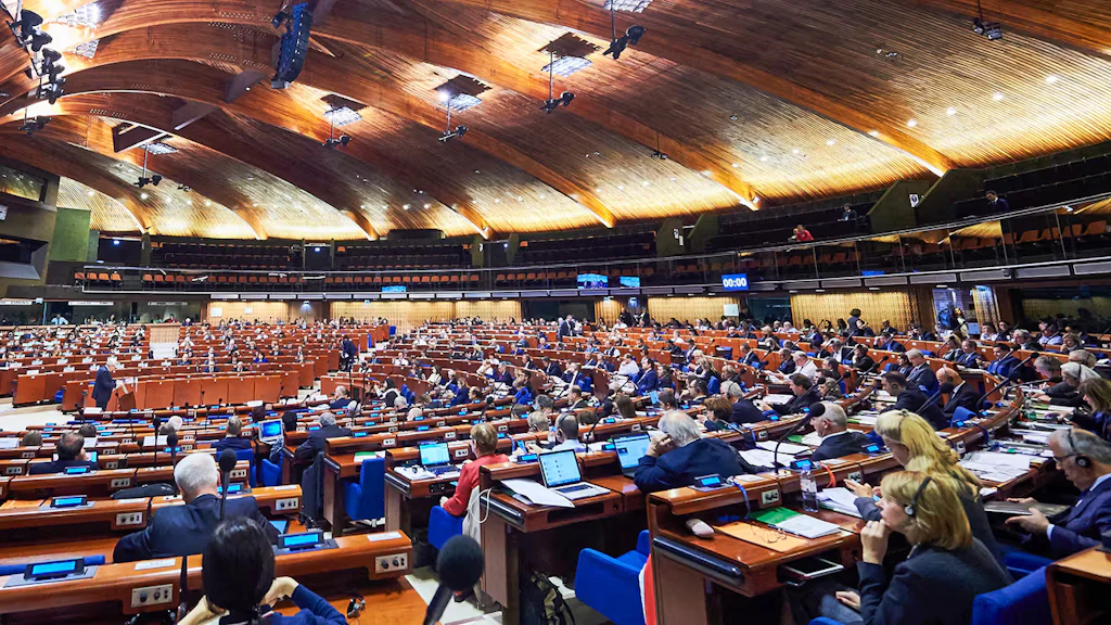 Una grande sala con un tetto in legno e file di sedili a semicerchio dove siedono i membri dell'Assemblea parlamentare del Consiglio d'Europa.