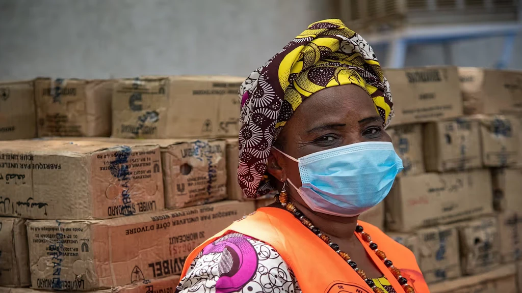 Una donna africana con una mascherina e un gilet arancione lavora in un deposito dove sono immagazzinate delle scatole.
